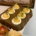 Close-up of garnished deviled eggs on a wooden platter with cheese and tomato slices.